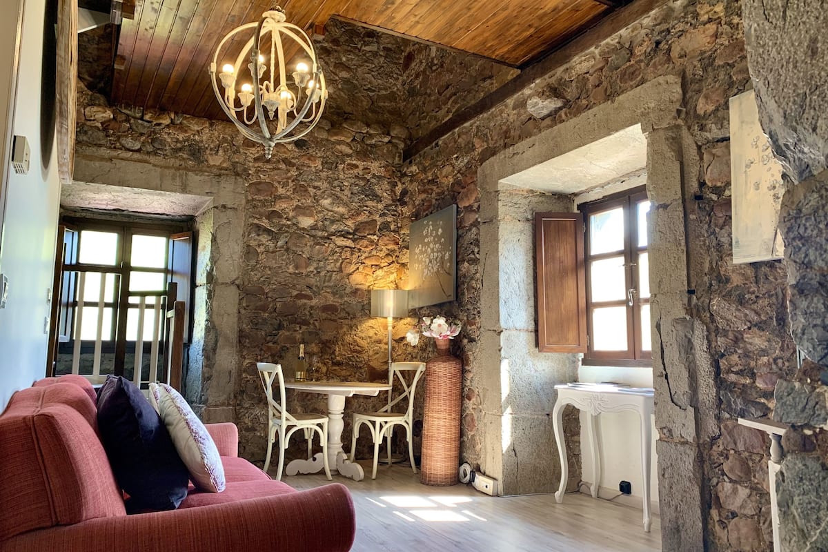 A rustic interior with exposed stone walls, a chandelier, a red sofa, and a dining area.