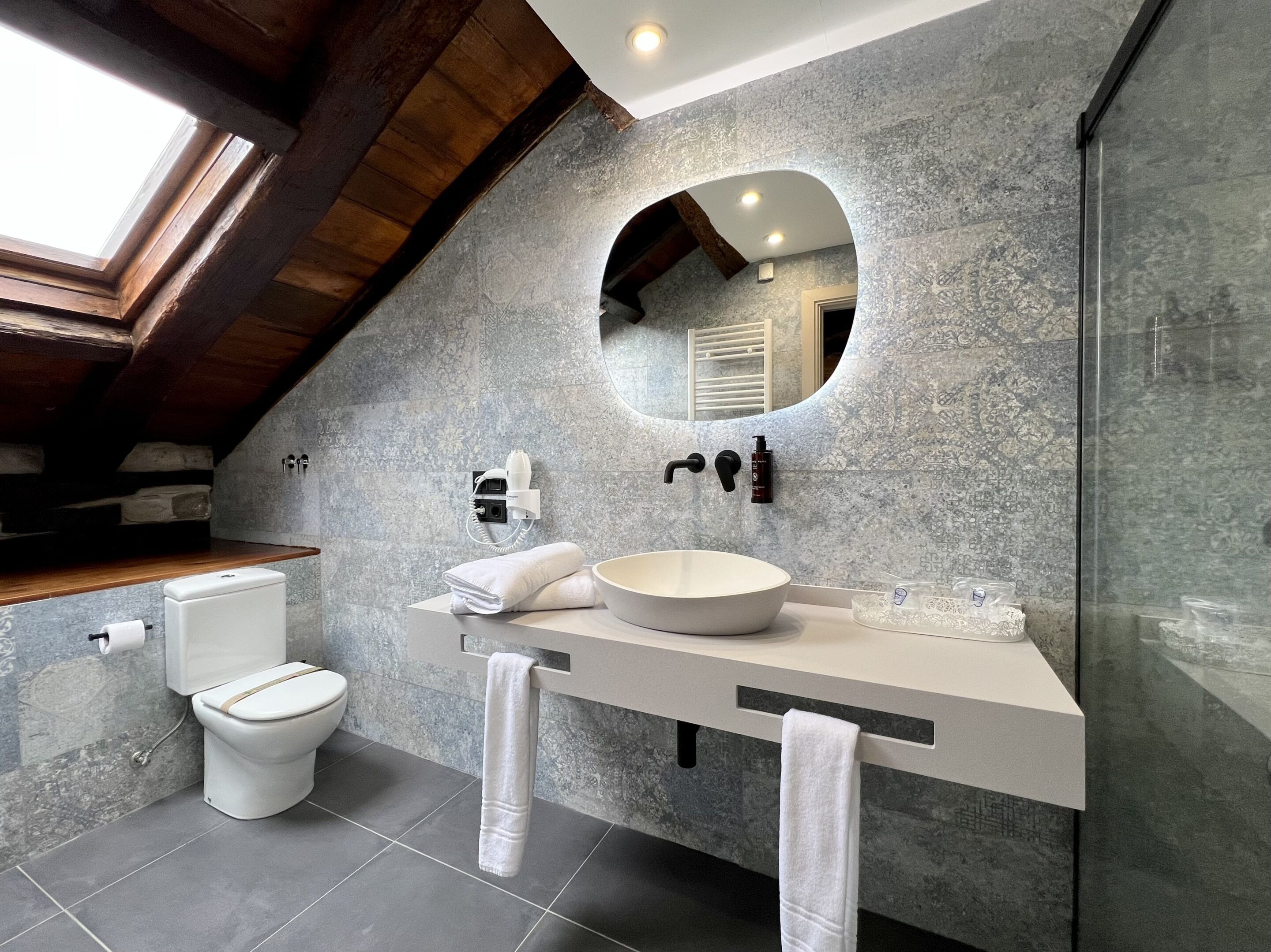 Un baño moderno con pared de piedra, espejo redondo, lavamanos blanco y retrete.