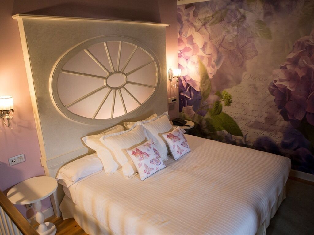 A bedroom with a large circular window, a bed with floral pillows, a nightstand, and a wall with a floral mural.