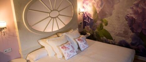 A bedroom with a large circular window, a bed with floral pillows, a nightstand, and a wall with a floral mural.