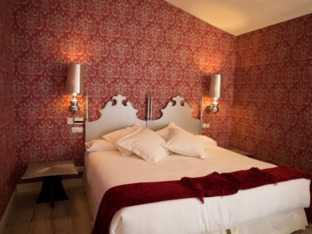 A neatly made bed with white linens and a red throw blanket, positioned in a room with patterned wallpaper and two wall-mounted lamps.