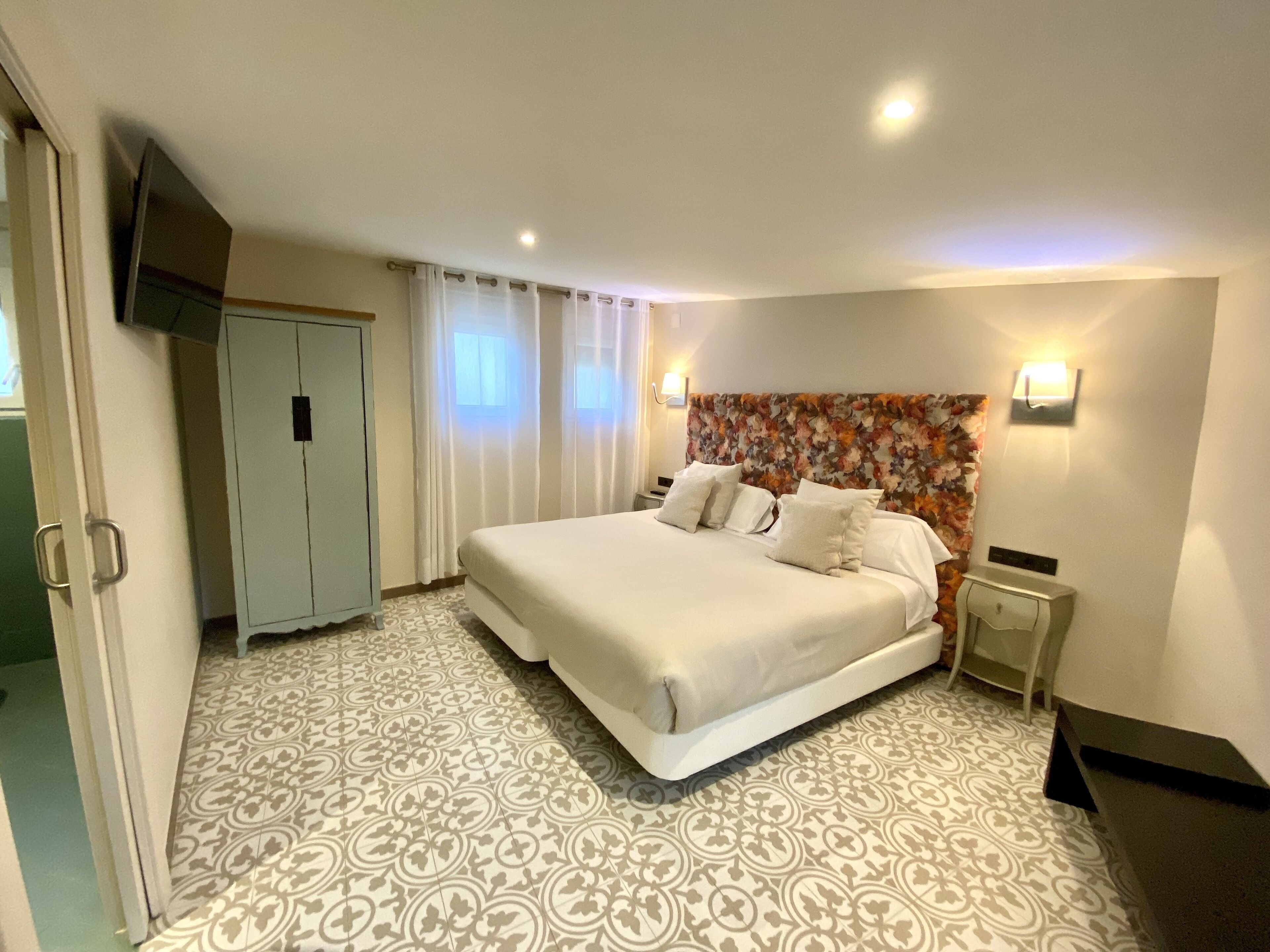 A hotel room with a large bed, a TV mounted on the wall, a wardrobe, and a patterned floor.