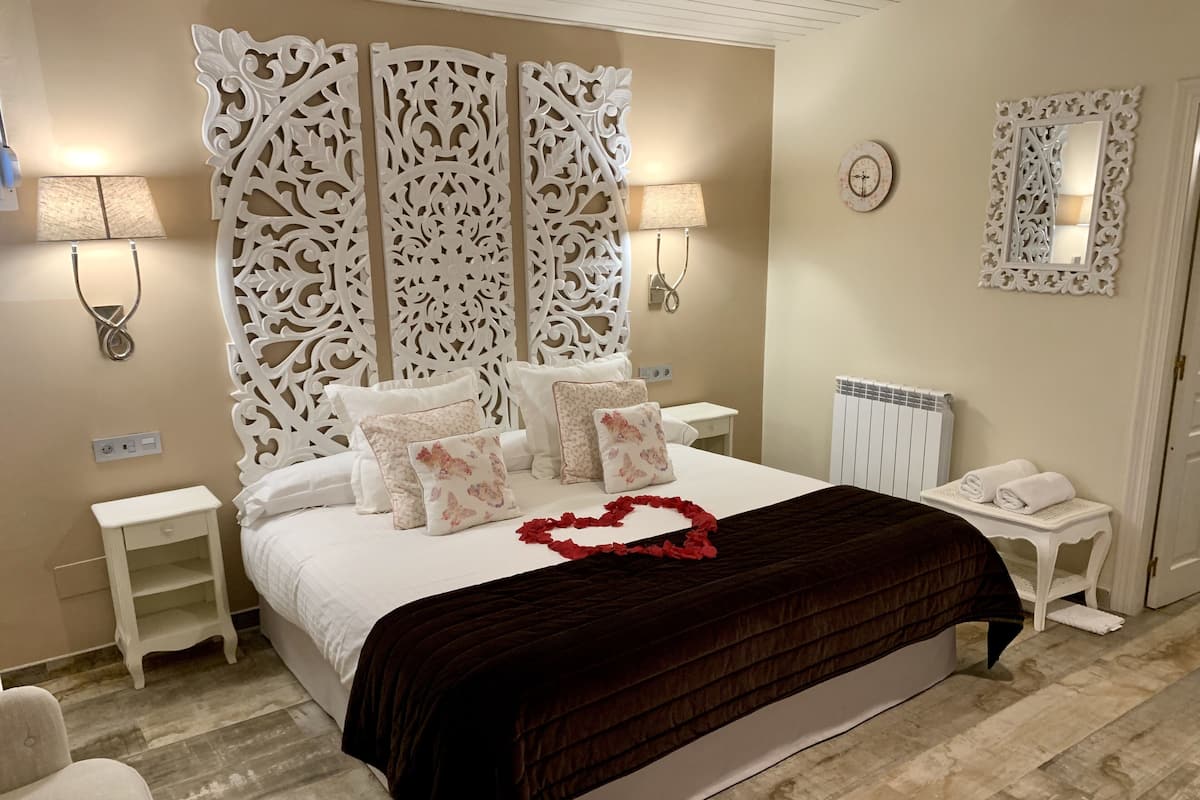 A bedroom with a large bed, white decorative headboard, two bedside lamps, a white nightstand, a white side table, and a mirror on the wall.