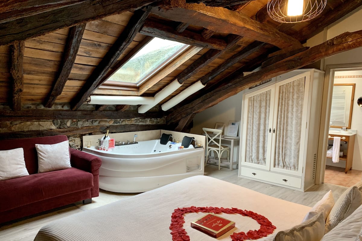 A cozy attic bedroom with a large bed, a red heart-shaped decoration, a white bathtub, and a wooden ceiling.