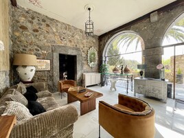 Amplia sala de estar con pared de piedra, chimenea y vista a palmeras a través de amplias ventanas arqueadas.