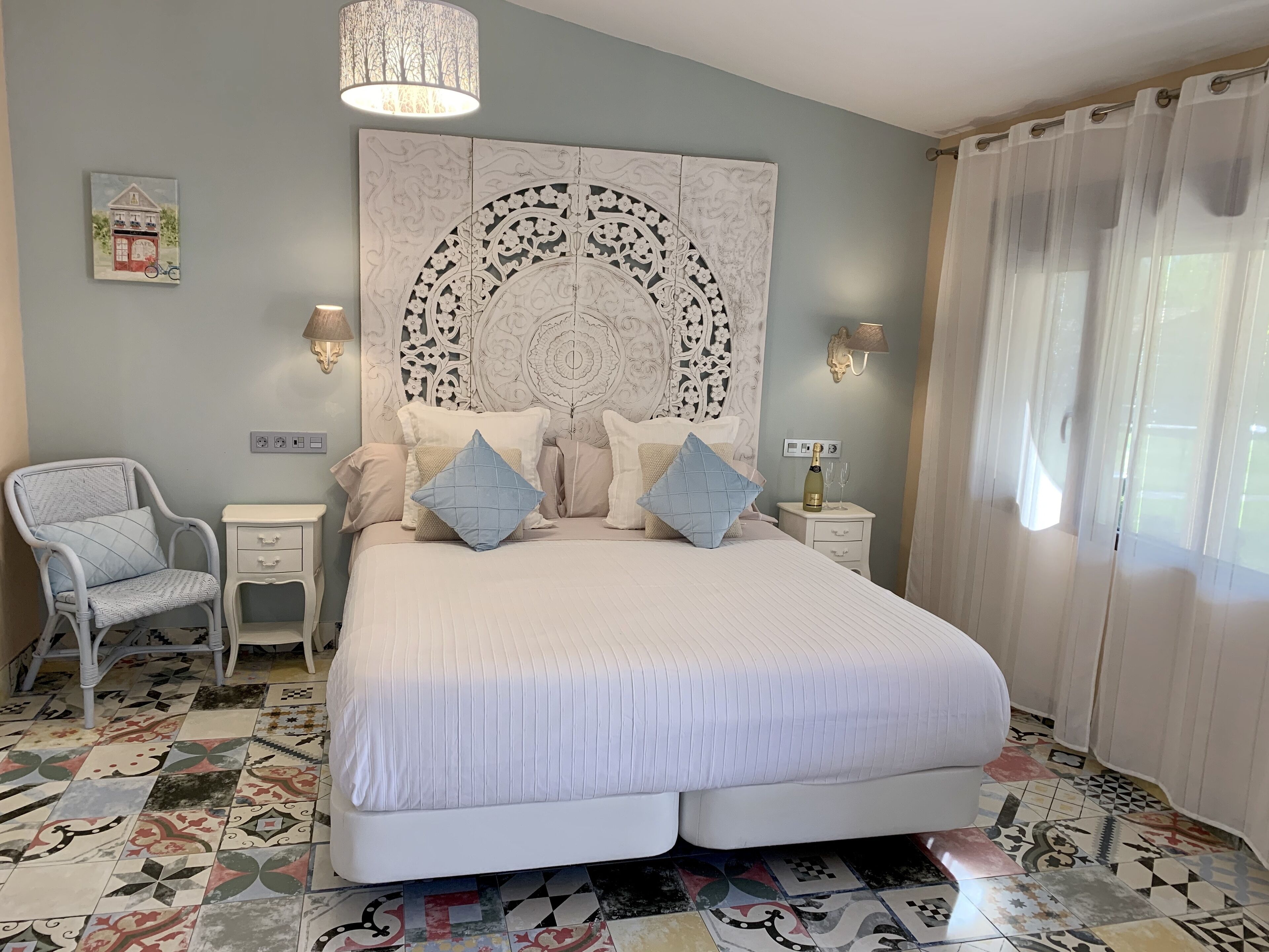 A bedroom with a large bed, a chair, a bedside table, a wall-mounted lamp, and a decorative panel above the bed.