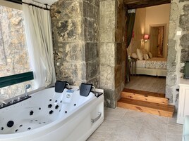 Un baño con una bañera grande, paredes de piedra y vistas hacia un dormitorio con cama y mesita de noche.