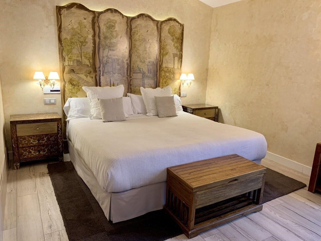 A bedroom with a large bed, two bedside tables with lamps, a decorative headboard, and a wooden bench.