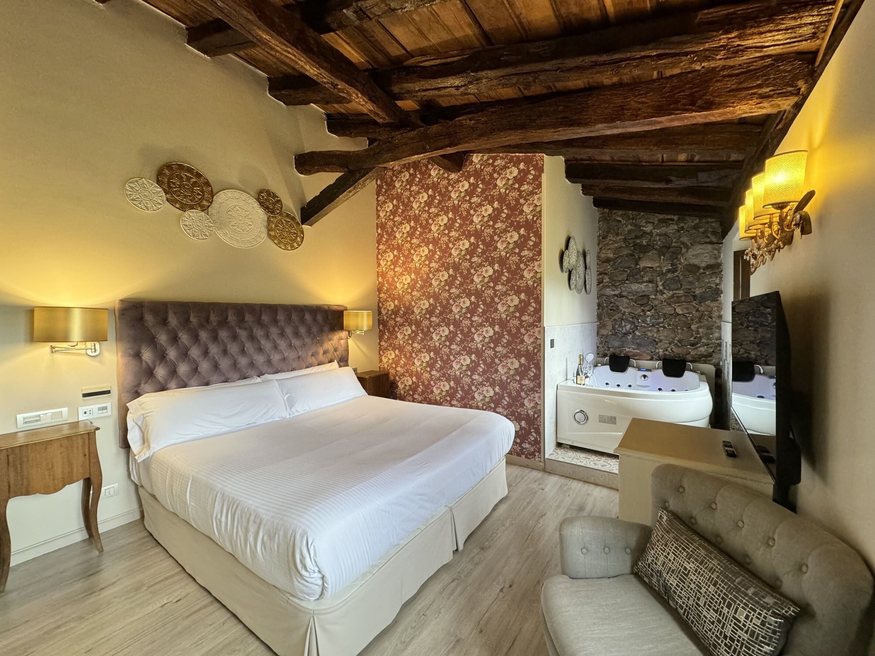 A bedroom with a large bed, a wooden ceiling, a stone wall, and a spa area with a bathtub.