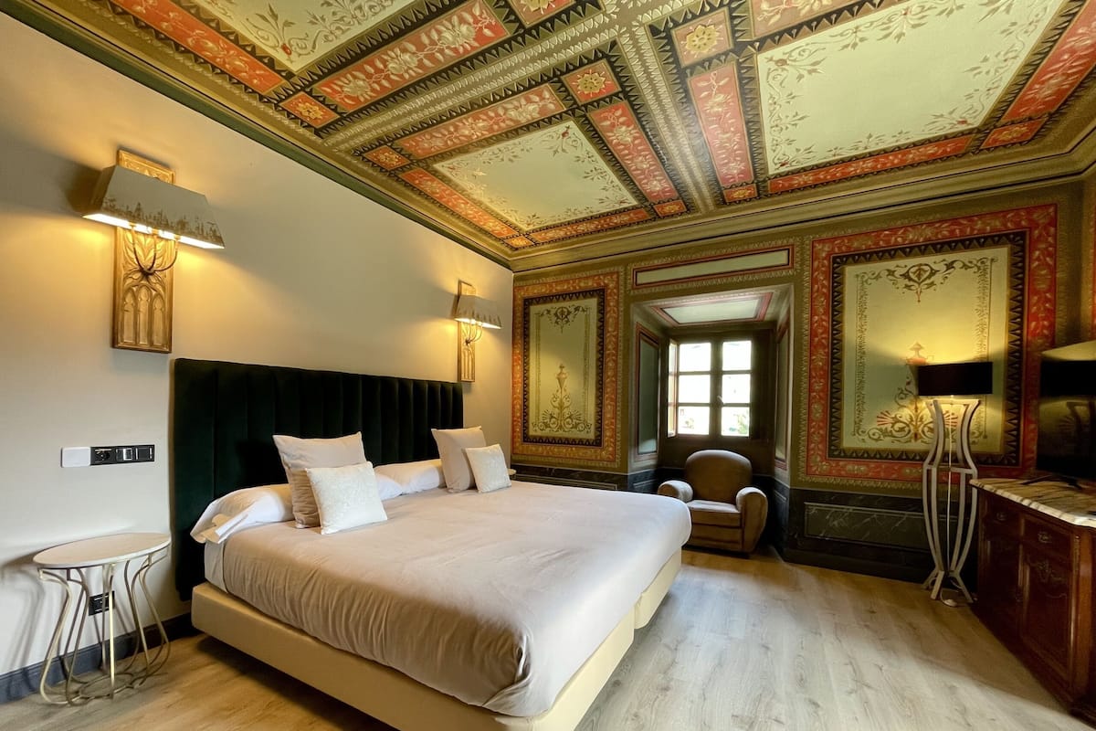 A spacious bedroom with a large bed, a small round table, and a chair. The room features ornate ceiling decorations and a large window.