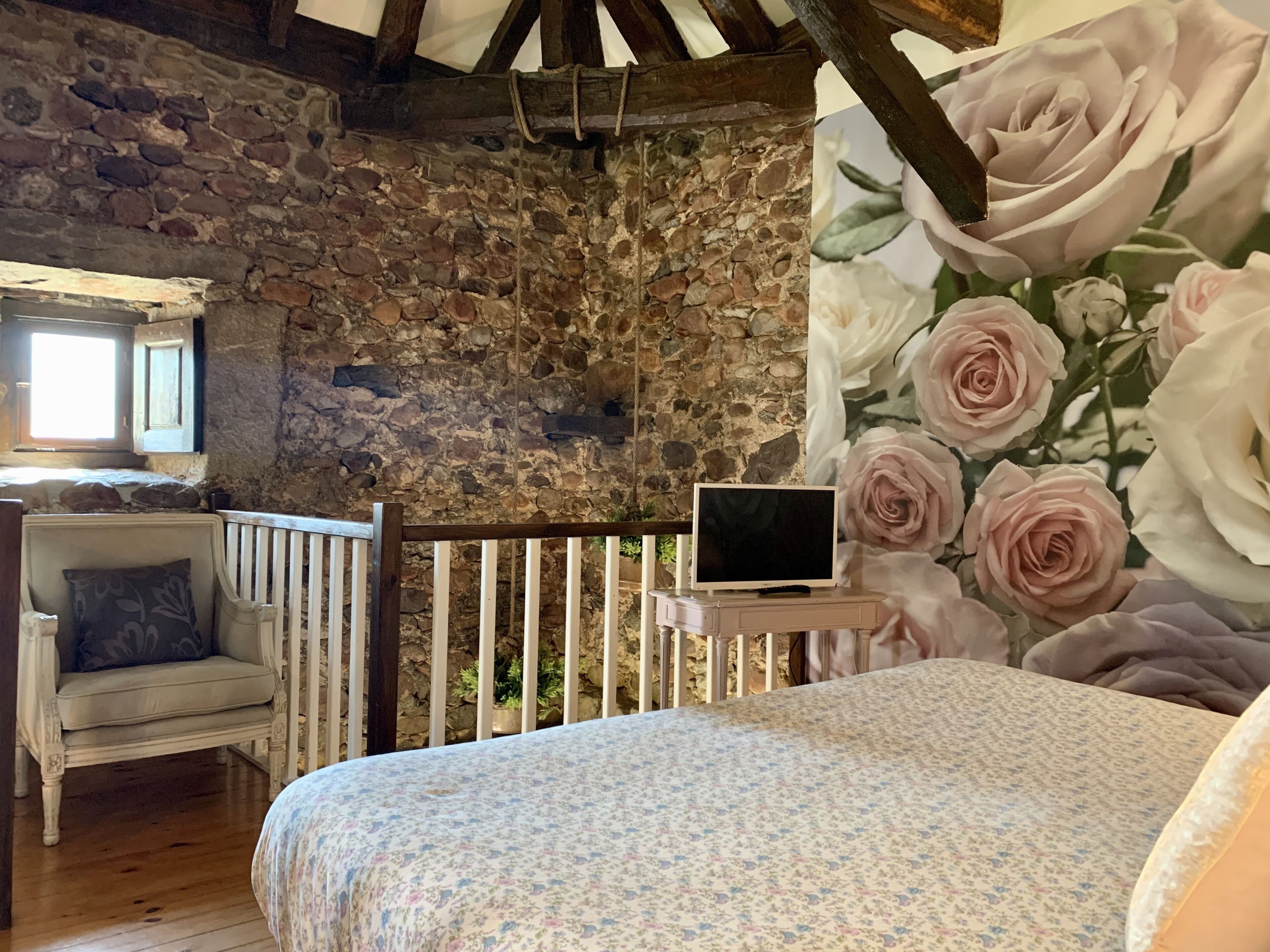 Una habitación rústica con pared de piedra, una cama de madera, una silla blanca y un televisor sobre una mesita.