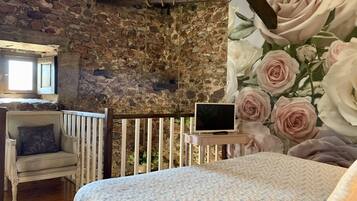 Una habitación rústica con pared de piedra, una cama de madera, una silla blanca y un televisor sobre una mesita.
