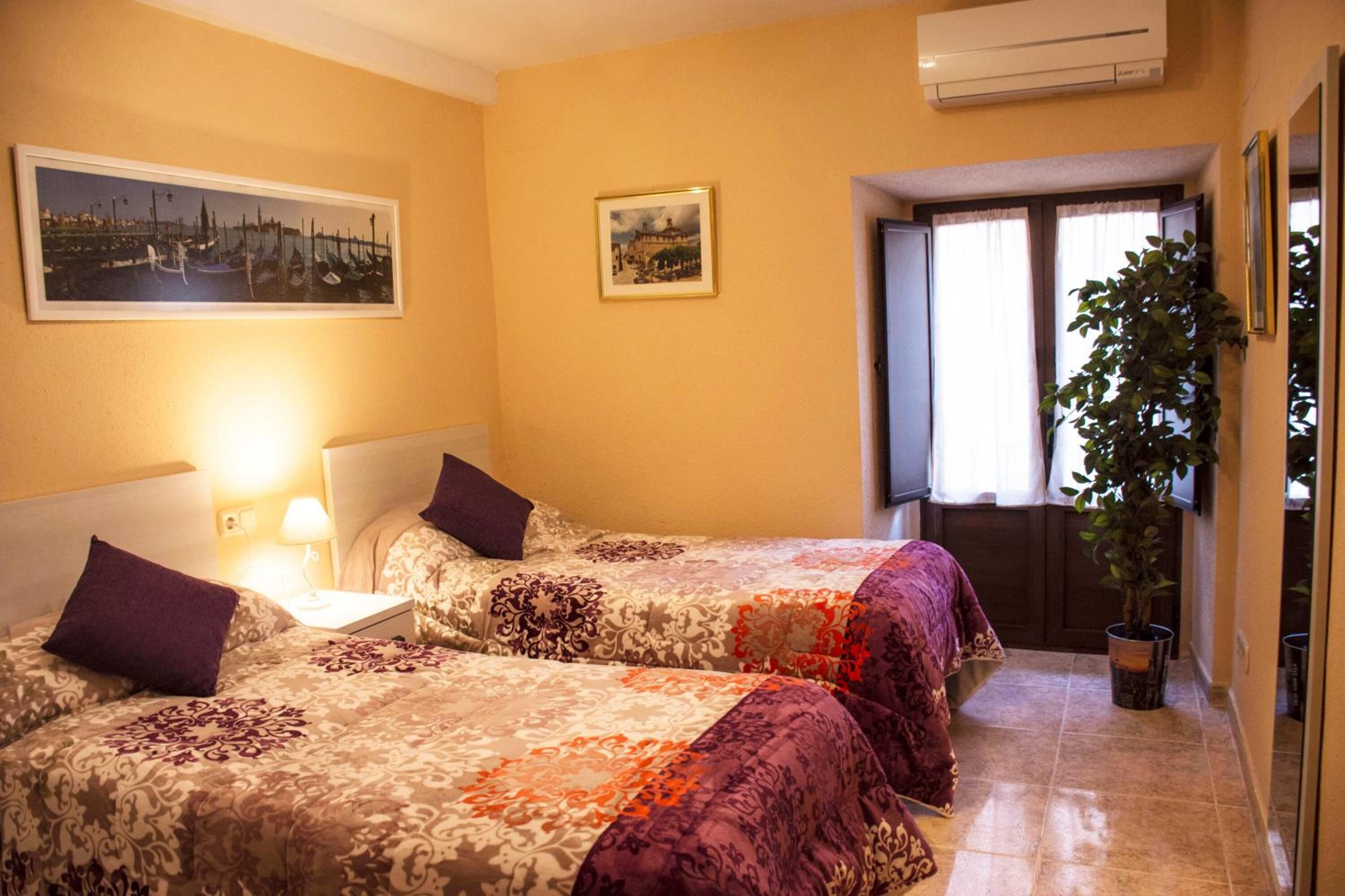 Habitación de hotel con dos camas, una mesita de noche, una lámpara, una planta en maceta y una ventana con cortinas.