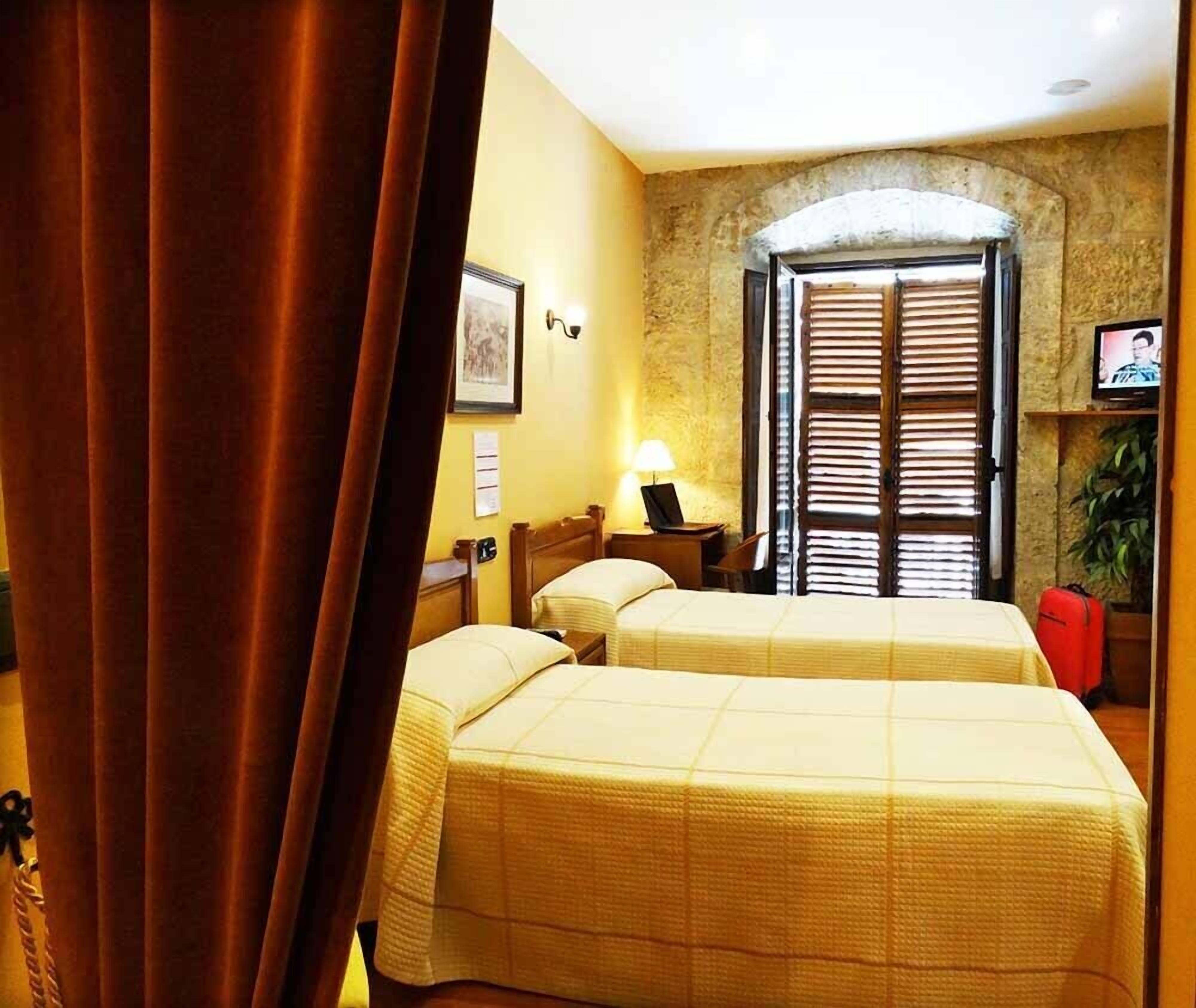 A hotel room with two beds, a lamp, a television, and a stone wall.