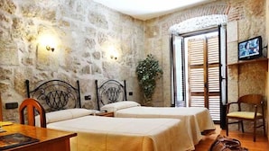 A room with two beds, a desk, and a chair. There is a stone wall, a plant, and a TV on a shelf. - Palacio Antigua Audiencia (Ciudad Rodrigo)