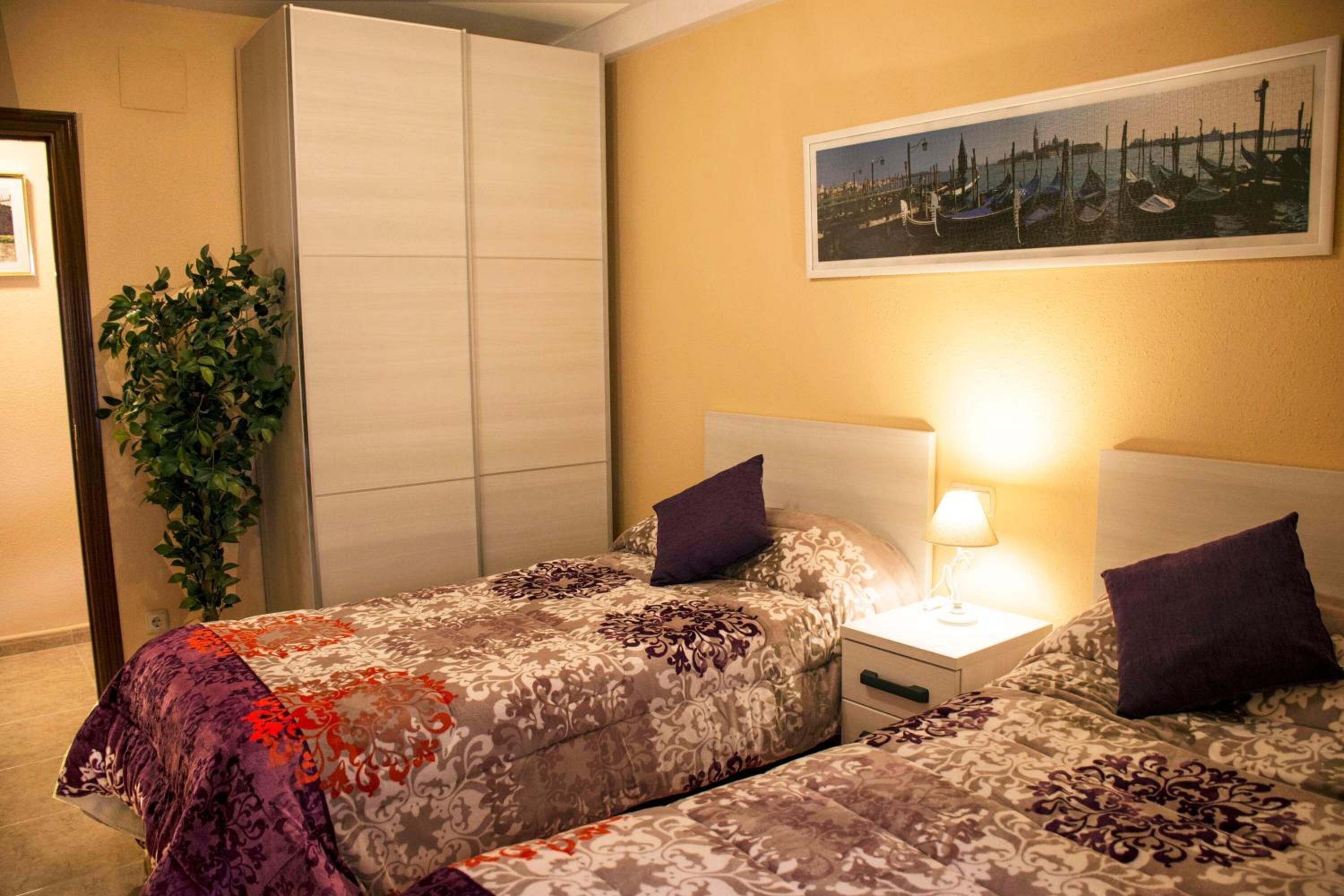 Une chambre d’hôtel avec deux lits, une table de chevet avec une lampe, une grande plante en pot et un tableau encadré au mur.