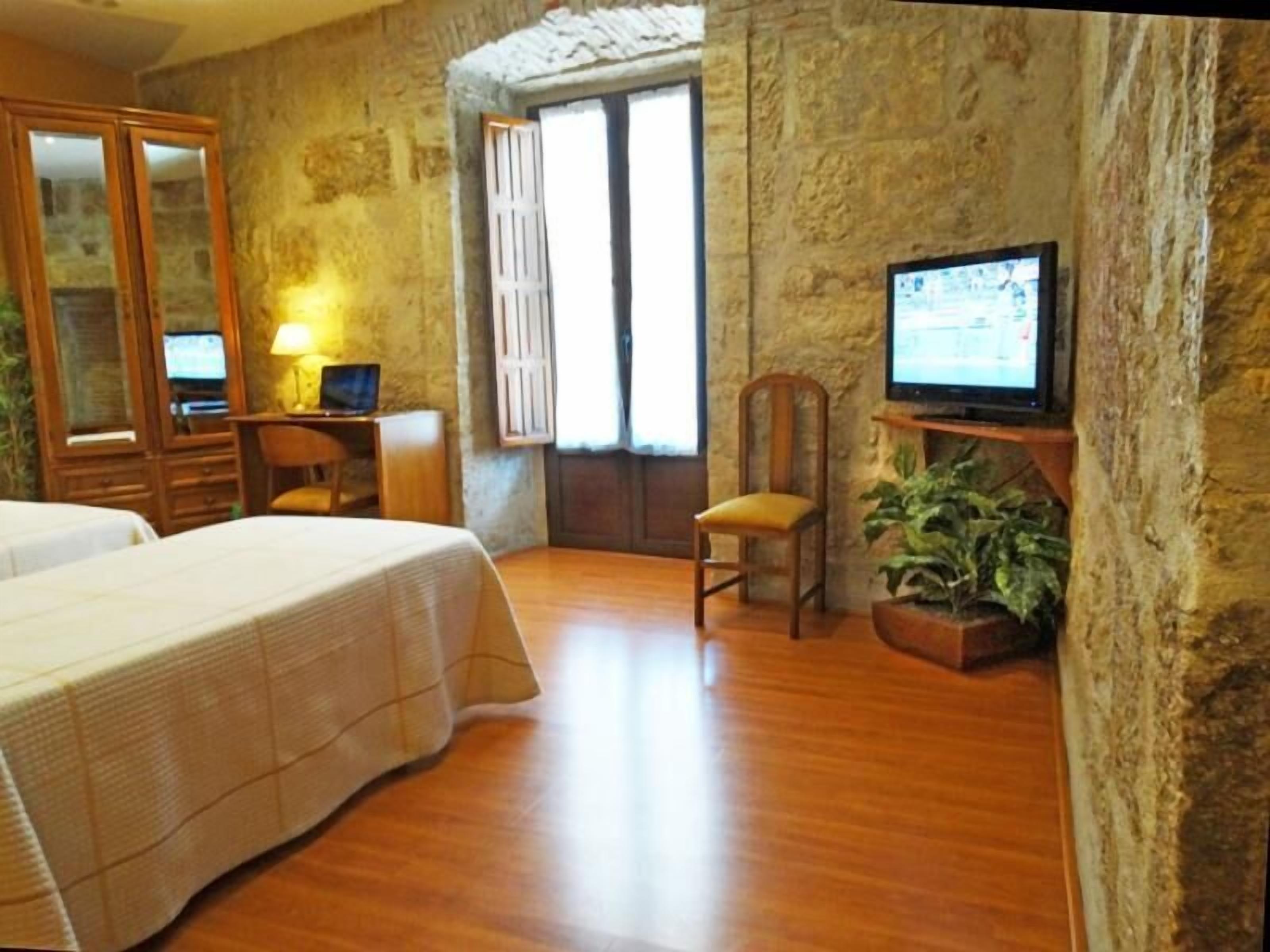 Habitación de hotel con cama, escritorio con computadora, televisor sobre un mueble bajo y ventana con cortinas.