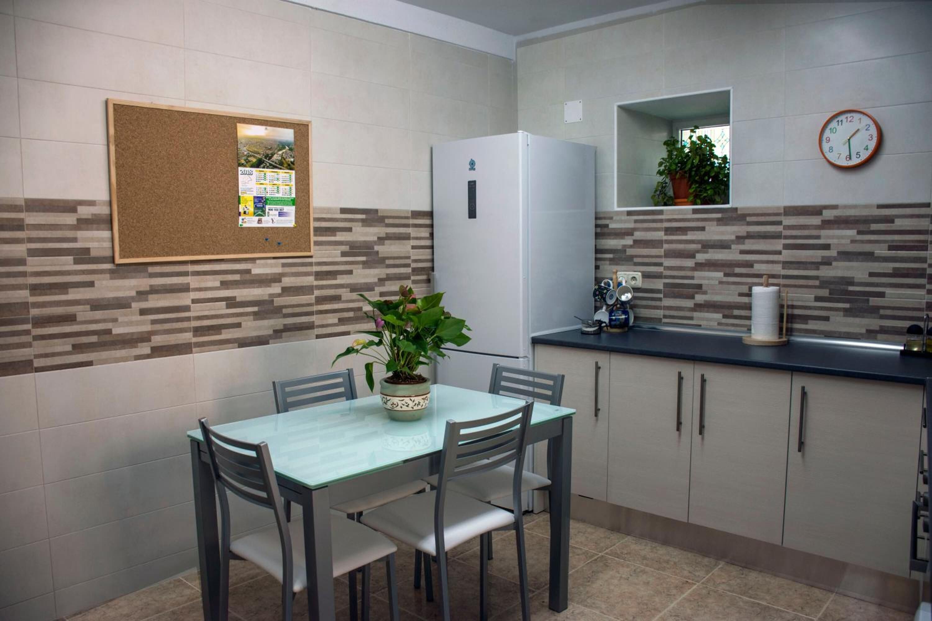 A compact kitchen with a dining table, chairs, a refrigerator, a clock, and a bulletin board.