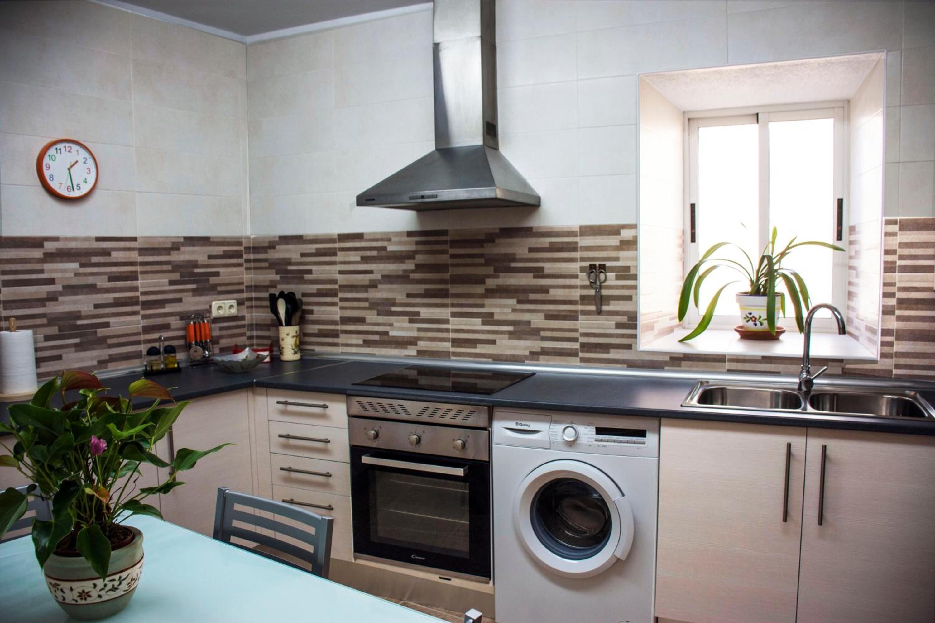 A compact kitchen with a built-in oven, a washing machine, a stainless steel range hood, and a window with a potted plant.