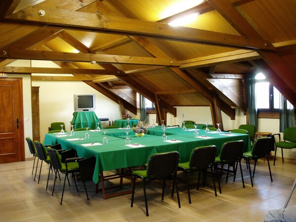Un salón de conferencias con techo alto y vigas de madera, equipado con mesas y sillas, una televisión y una pequeña mesa con flores.