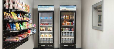 Dans un couloir, on trouve deux distributeurs automatiques de boissons en libre-service, avec des étagères remplies de collations sur le côté gauche.