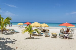 A beach with sun loungers and umbrellas, palm trees, and a clear blue sky.