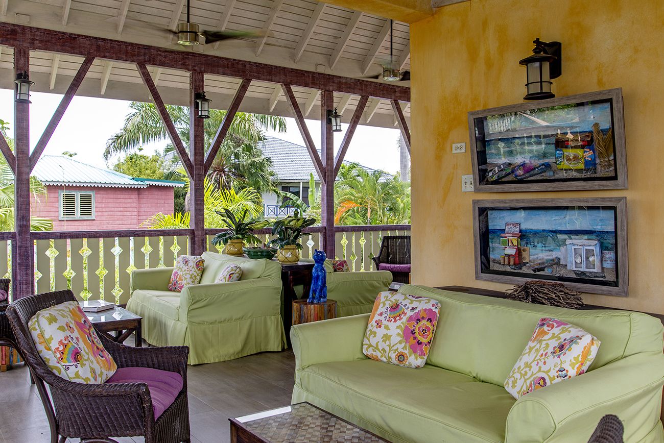 A covered outdoor seating area with wicker furniture and cushions, a wooden table, and a wall with framed artwork.