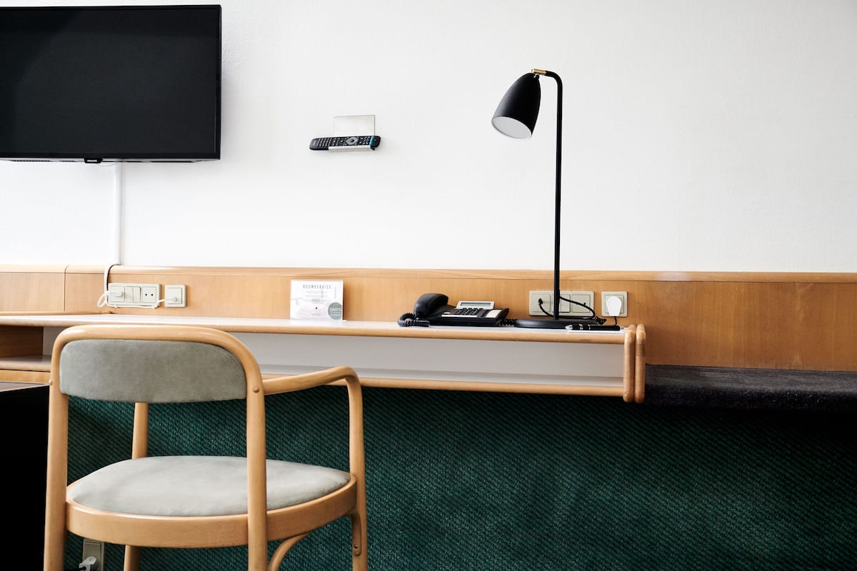 A hotel room with a wooden desk, a chair, a flat-screen TV mounted on the wall, a desk lamp, and a telephone.