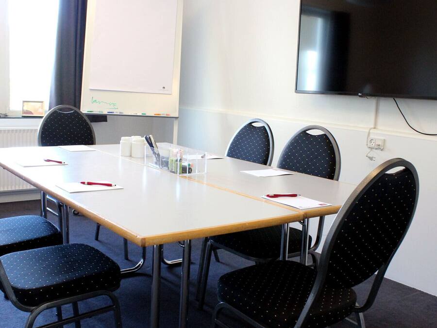 A conference room with a long table, six chairs, a whiteboard, and a TV mounted on the wall.