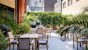 Un patio al aire libre con mesas y sillas de madera, rodeado de abundante vegetación y un muro de ladrillo.