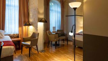 Une chambre d’hôtel avec un plancher en bois, un lit, deux fauteuils, un bureau avec une chaise et un lampadaire.