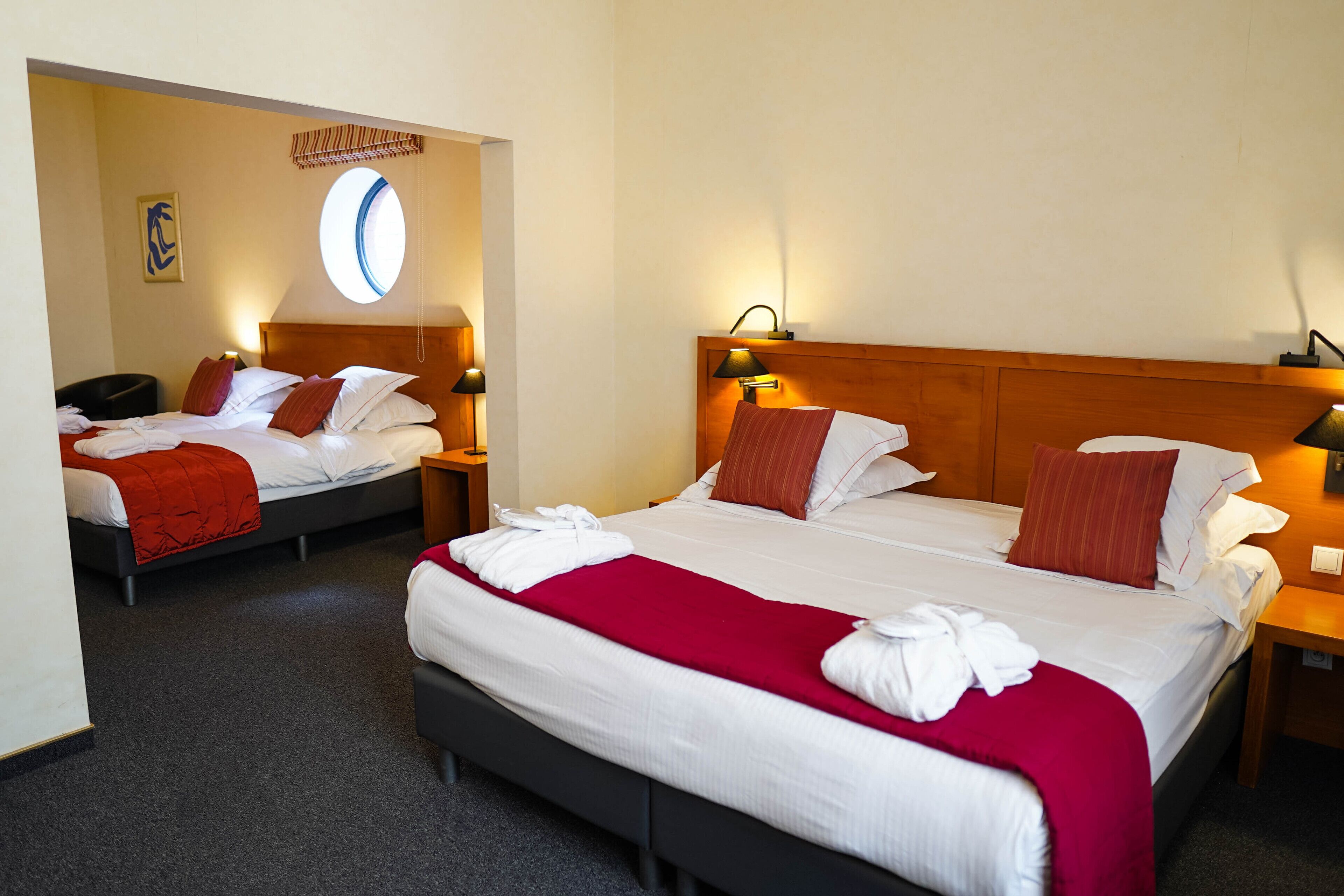 Een hotelkamer met twee bedden, elk met een houten hoofdbord, witte lakens en rode kussens. Er staan nachtlampjes en een klein tafeltje met een lamp.