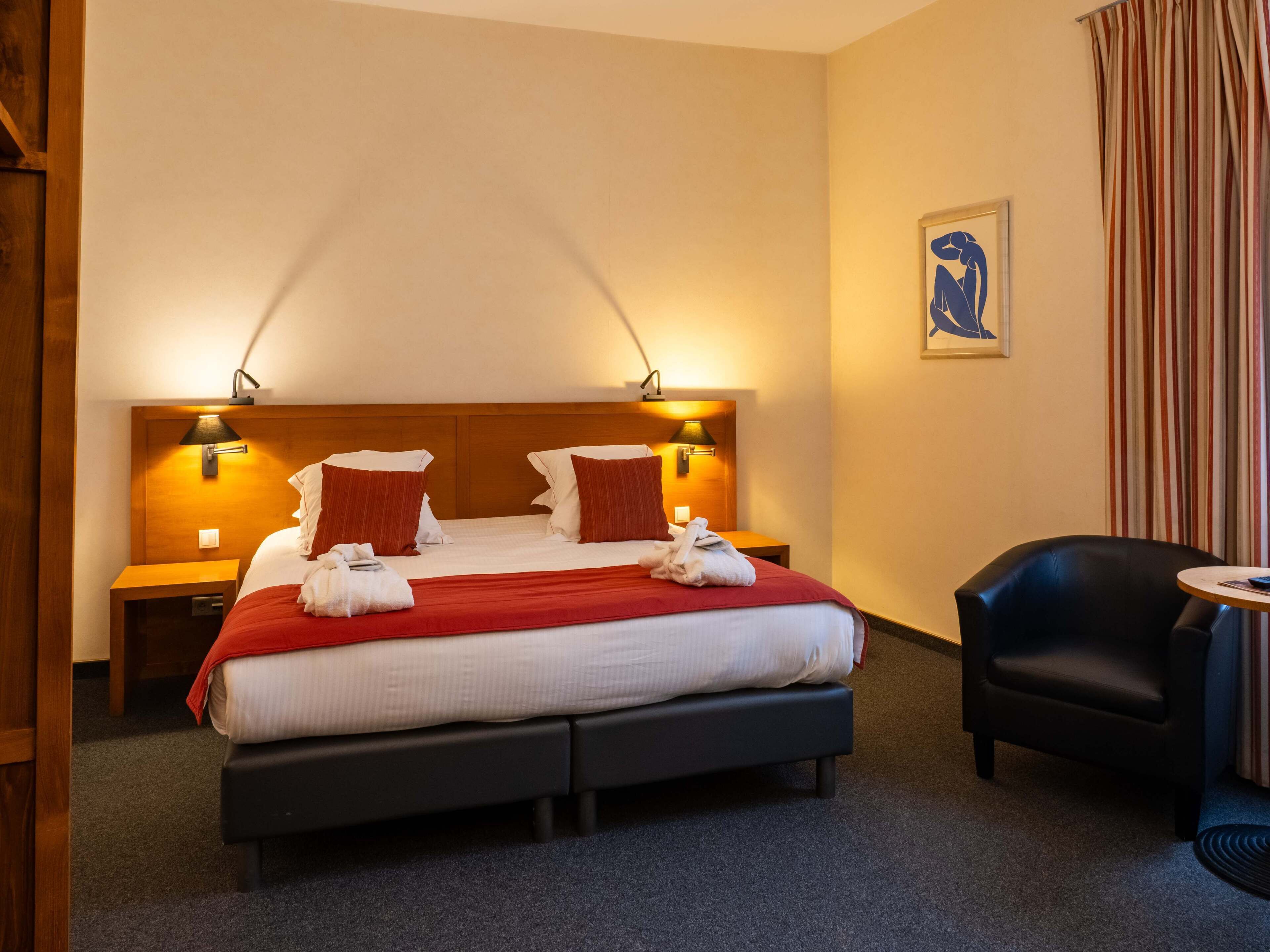 A hotel room with a large bed, two bedside tables, a chair, a small table, and a painting on the wall.