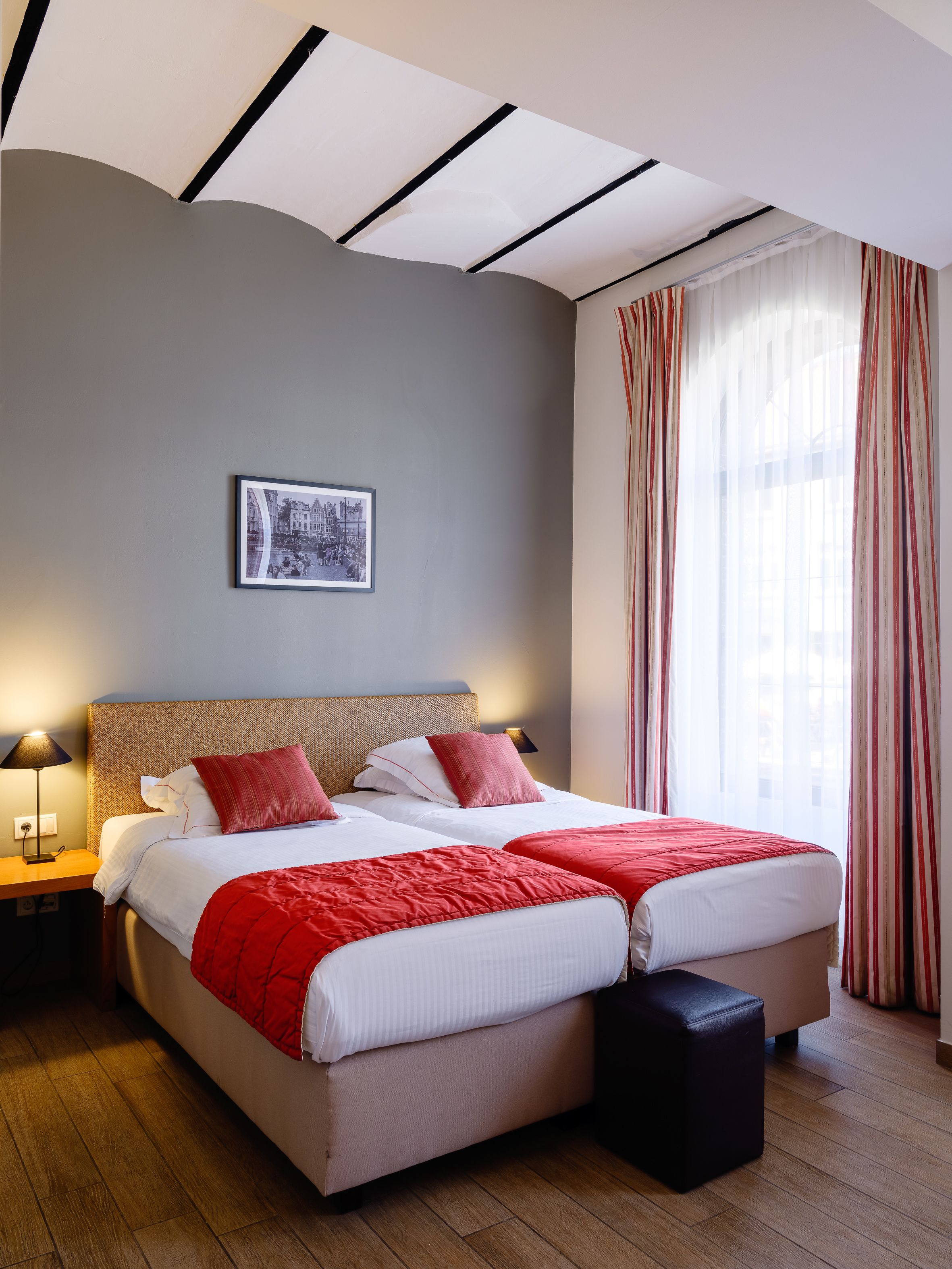 Un lit double avec une literie rouge et blanche, une table de chevet avec une lampe, un tableau encadré au mur et une fenêtre avec des rideaux à rayures.
