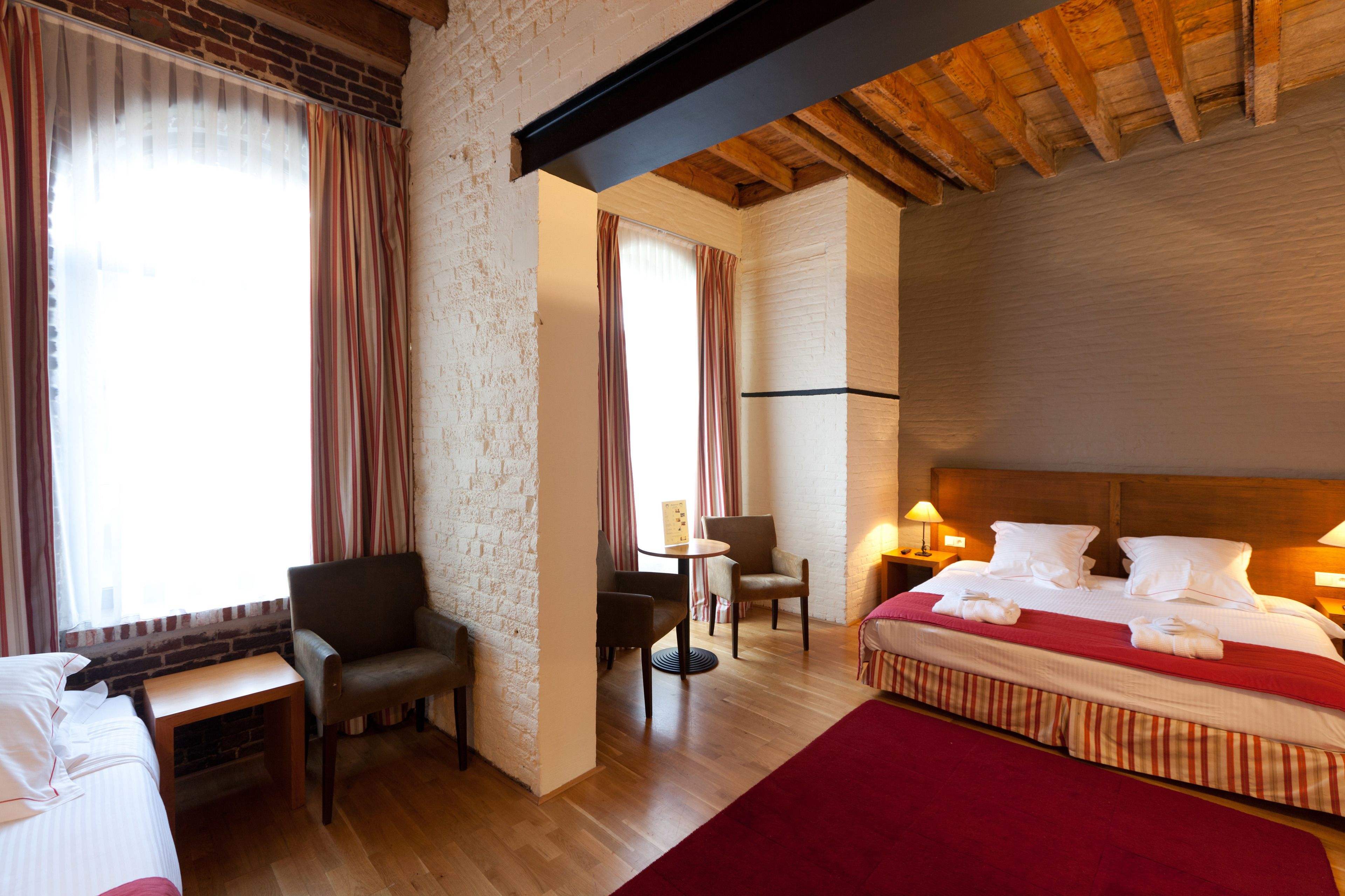 Een hotelkamer met een houten plafond, een bed met witte lakens, twee stoelen, een kleine tafel en een raam met gordijnen.