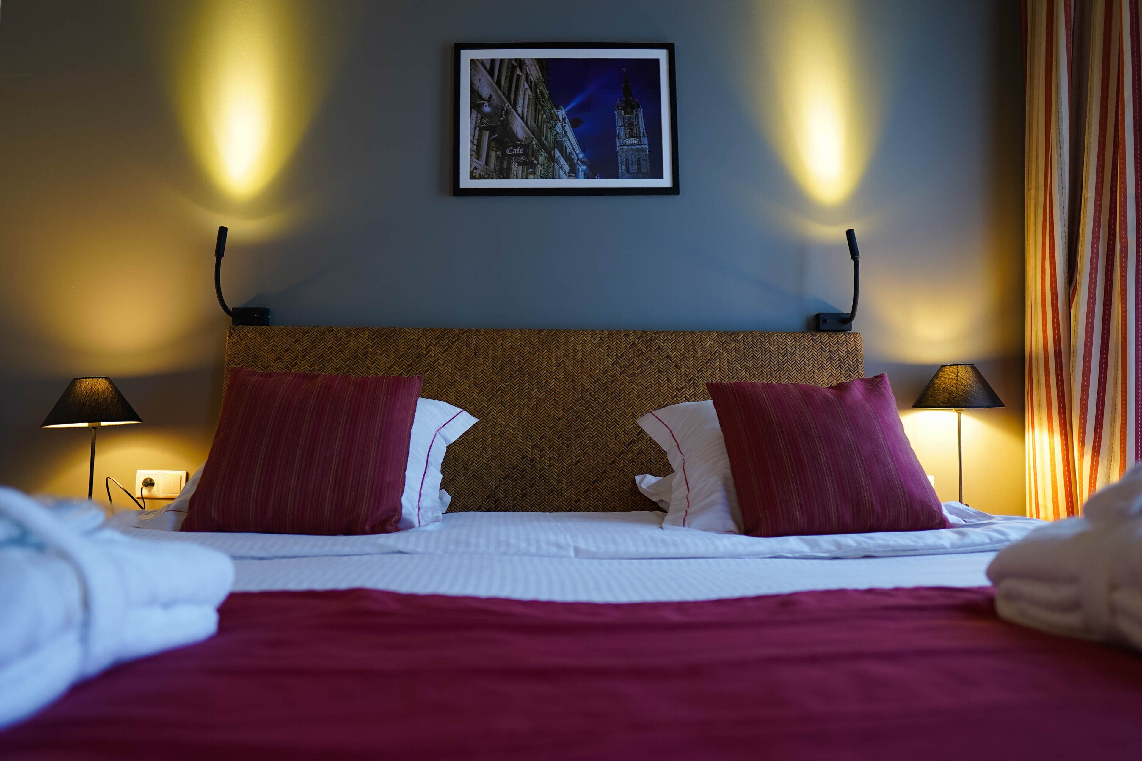 A hotel room with a bed, two bedside lamps, a framed picture on the wall, and a window with curtains.