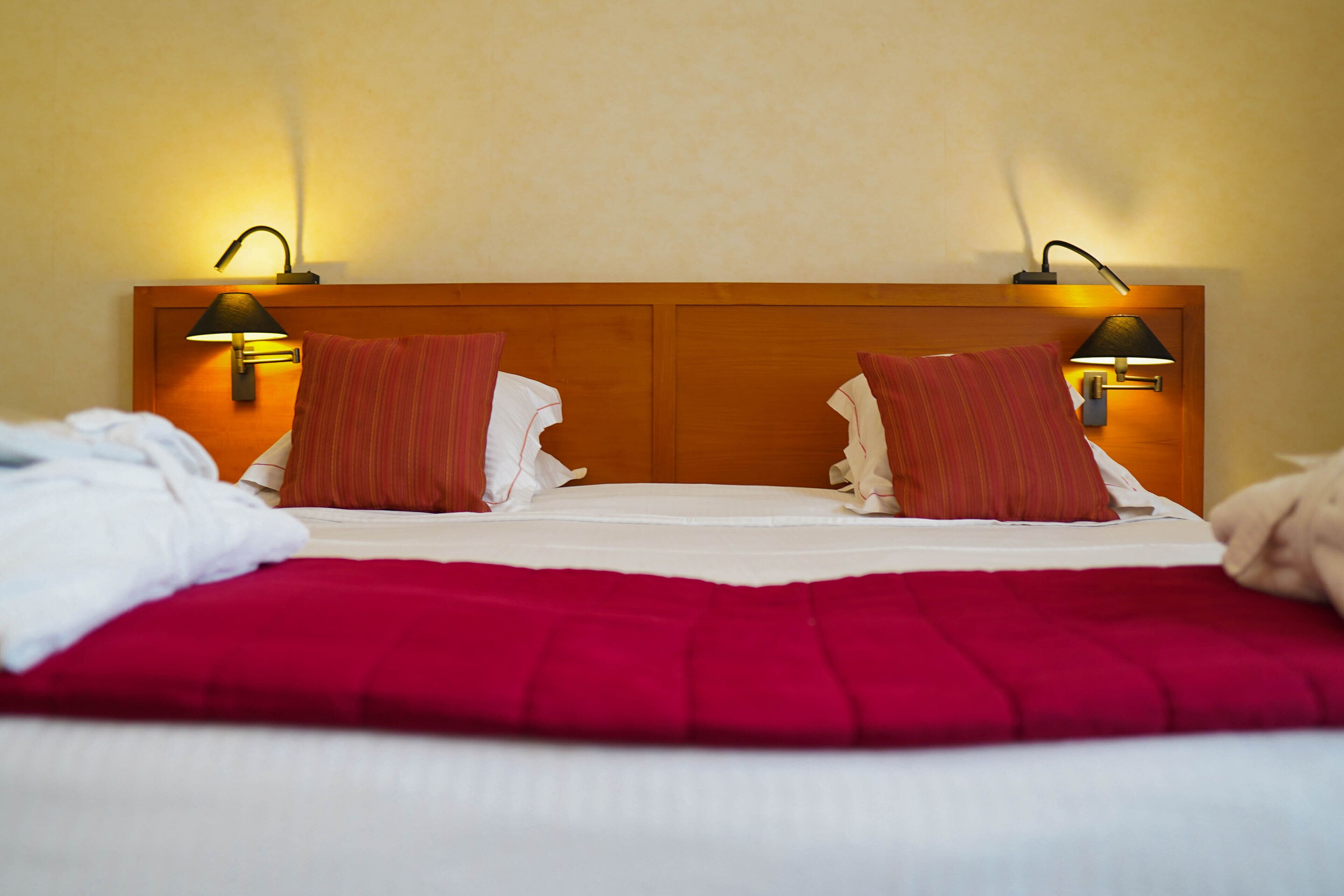 Une chambre d’hôtel avec une tête de lit en bois, deux lampes de chevet, un plaid rouge et une couvre-lit blanche.