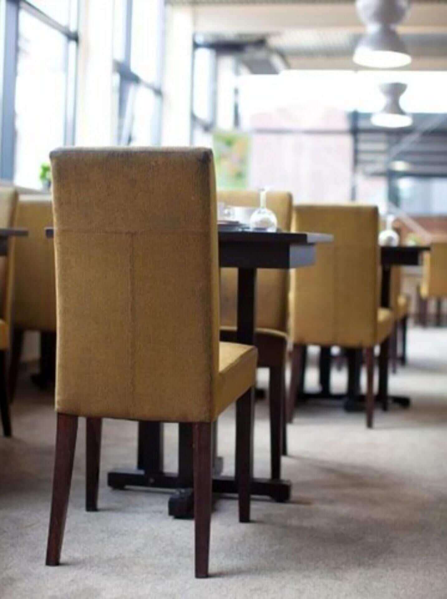 A row of dining tables with chairs, a carpeted floor, and large windows in the background.