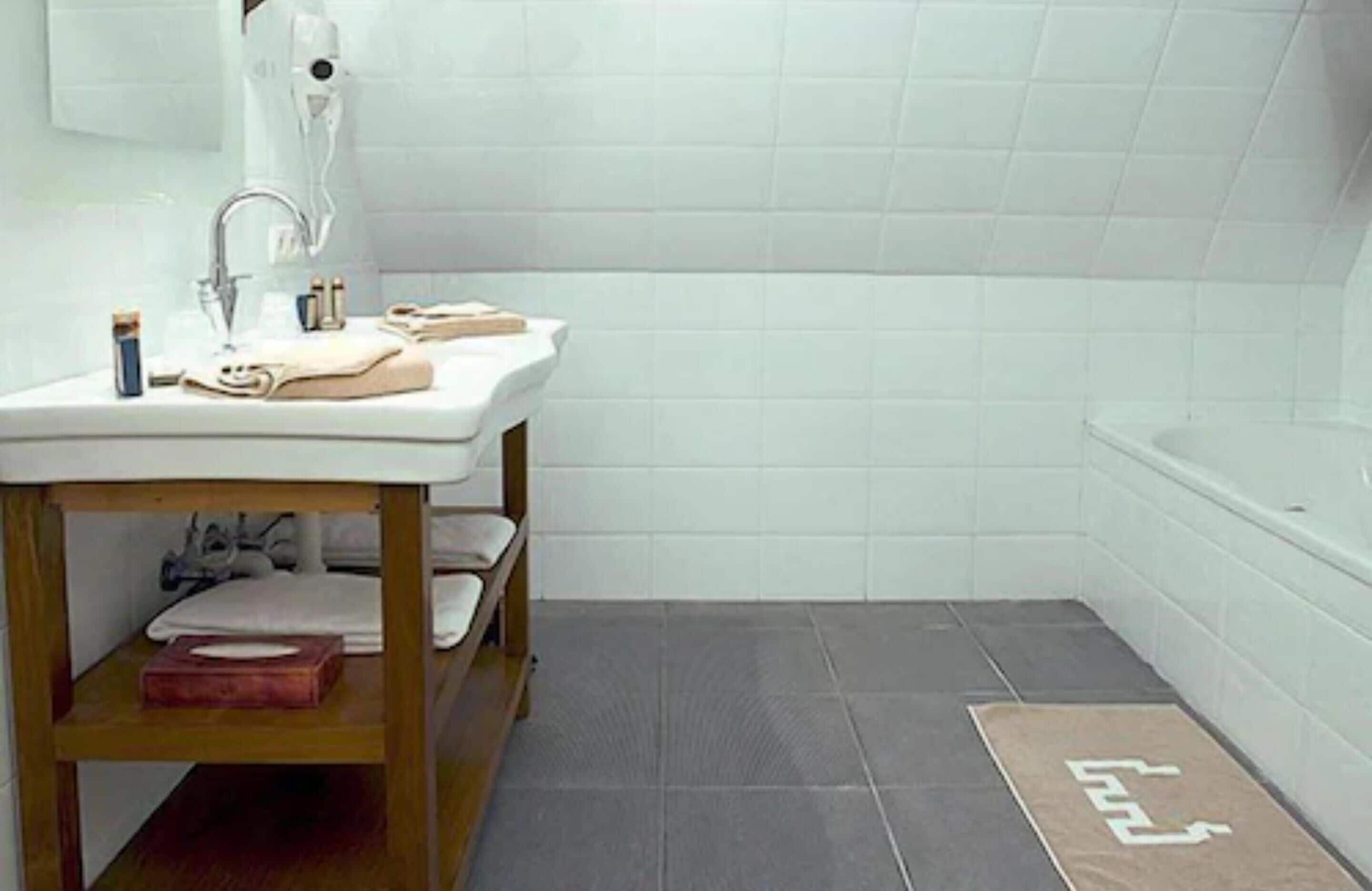 Une salle de bain avec un lavabo blanc, une étagère en bois, une baignoire et un sol carrelé foncé.