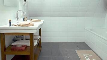 Une salle de bain avec un lavabo blanc, une étagère en bois, une baignoire et un sol carrelé foncé.