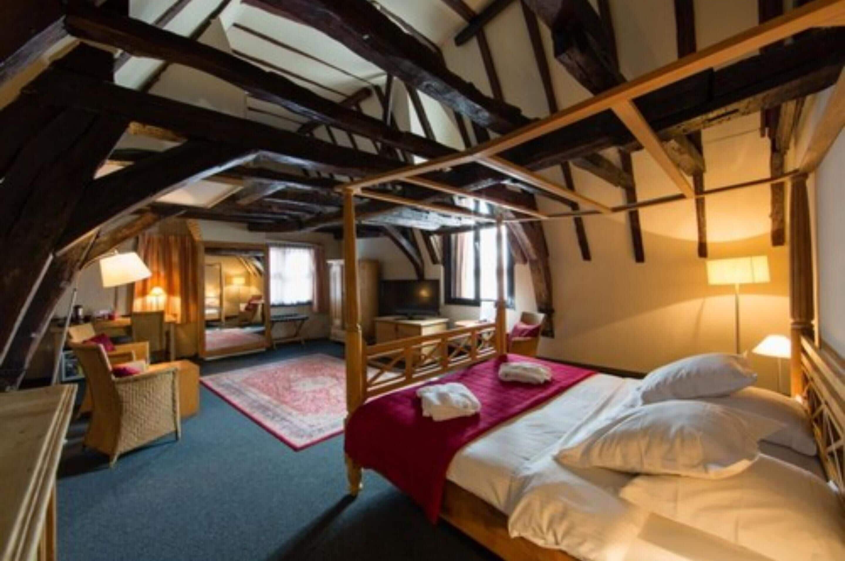 A room with a four-poster bed, wooden beams, a desk with a chair, and a window.