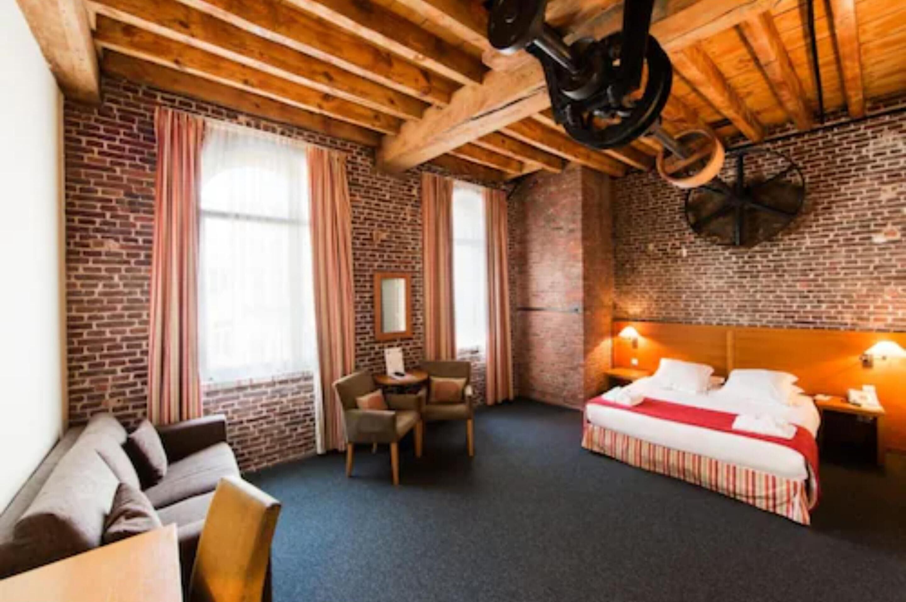 Habitación de hotel con vigas de madera a la vista, paredes de ladrillo, cama con colcha a rayas rojas y blancas, una pequeña mesa con sillas y una ventana con cortinas.