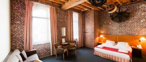 Habitación de hotel con vigas de madera a la vista, paredes de ladrillo, cama con colcha a rayas rojas y blancas, una pequeña mesa con sillas y una ventana con cortinas.