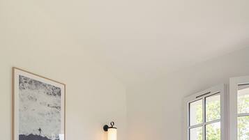 A bedroom with a bed, a ceiling fan, a wall-mounted light, a framed picture, and a window with a view of greenery.