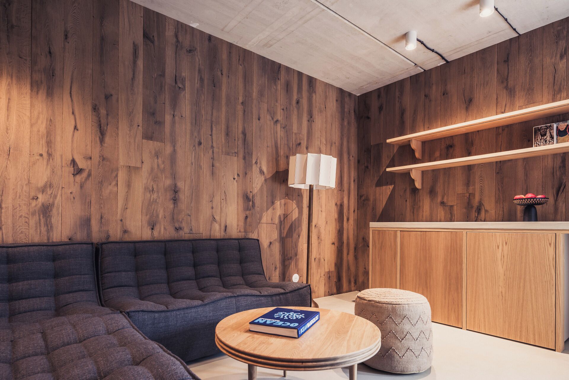 Una habitación moderna con una pared de madera, un sofá, una mesa redonda con un libro, una lámpara y un pequeño otoman.