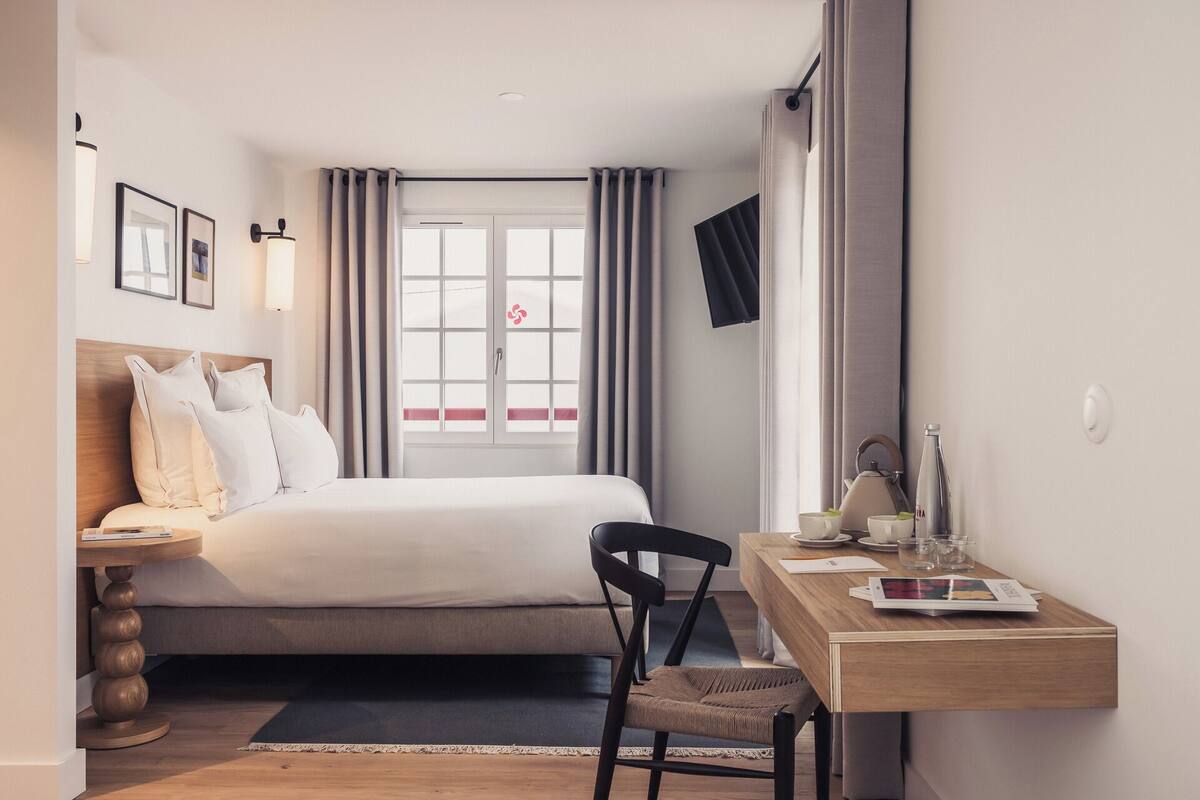 Une chambre d’hôtel comprenant un lit, un bureau, une chaise, un téléviseur fixé au mur et une fenêtre avec des rideaux.