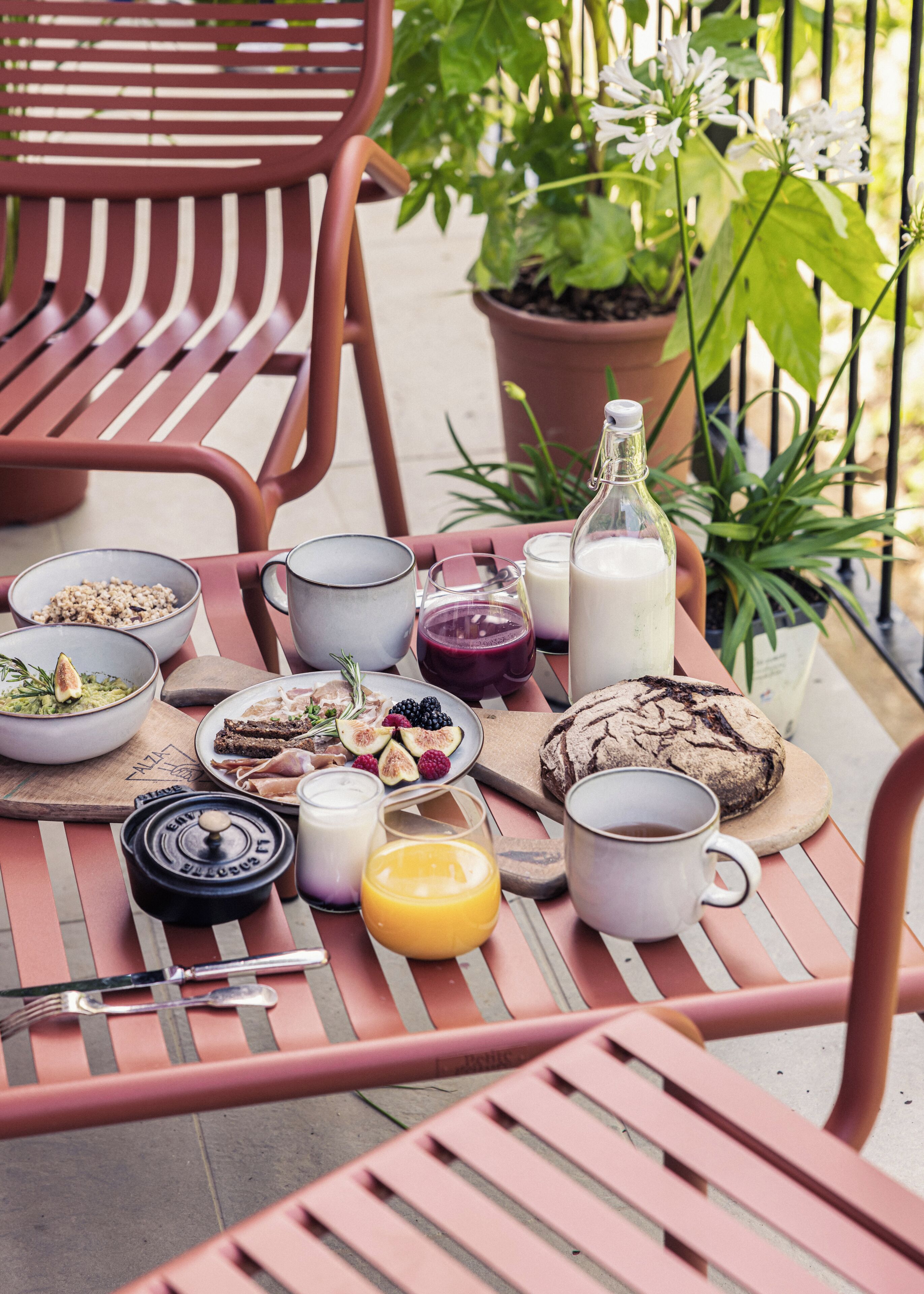 A table set with breakfast items including cereal, fruit, bread, and beverages, with a potted plant in the background.