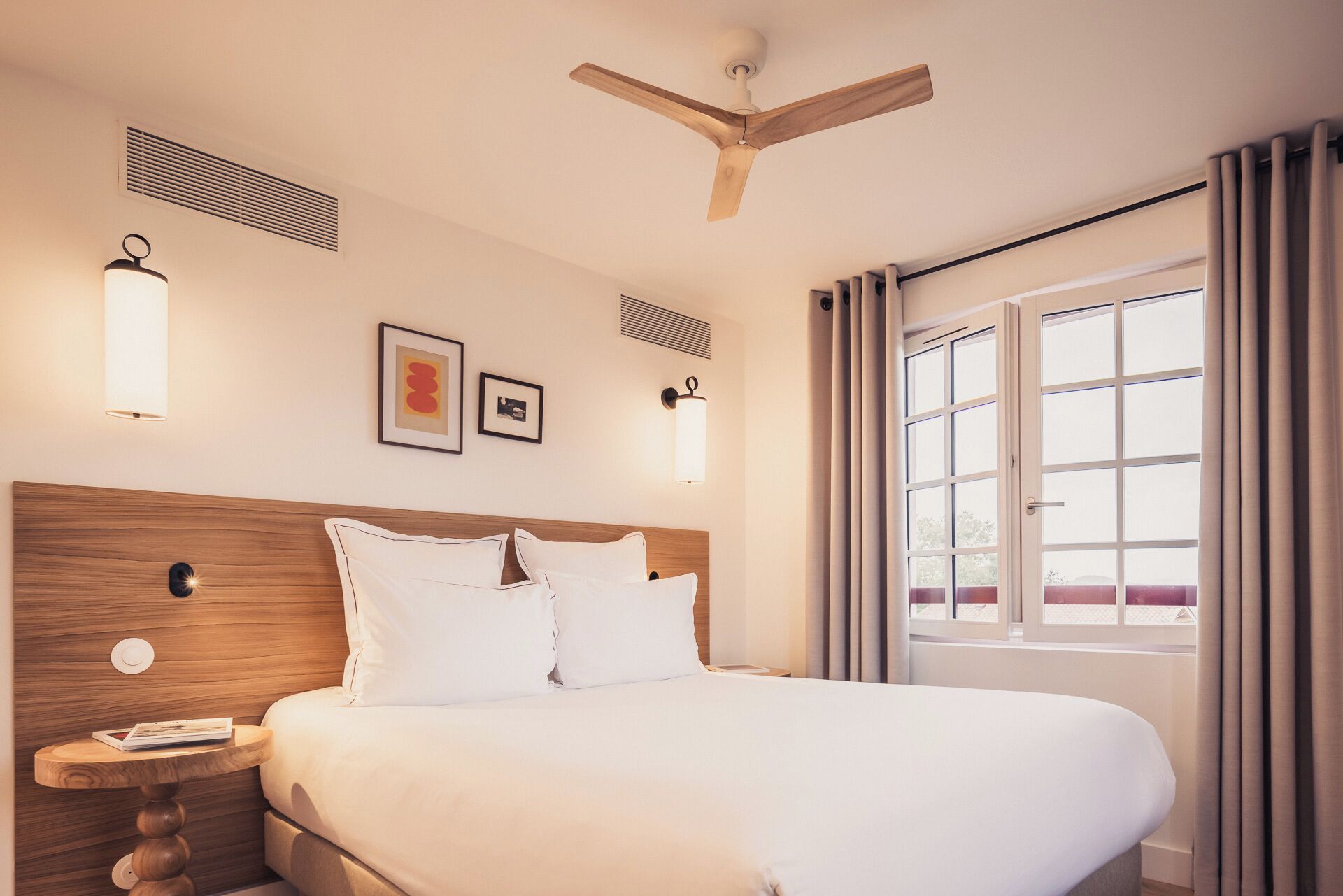 A hotel room with a wooden headboard, a large bed, a bedside table with a lamp, a ceiling fan, and a window with curtains.