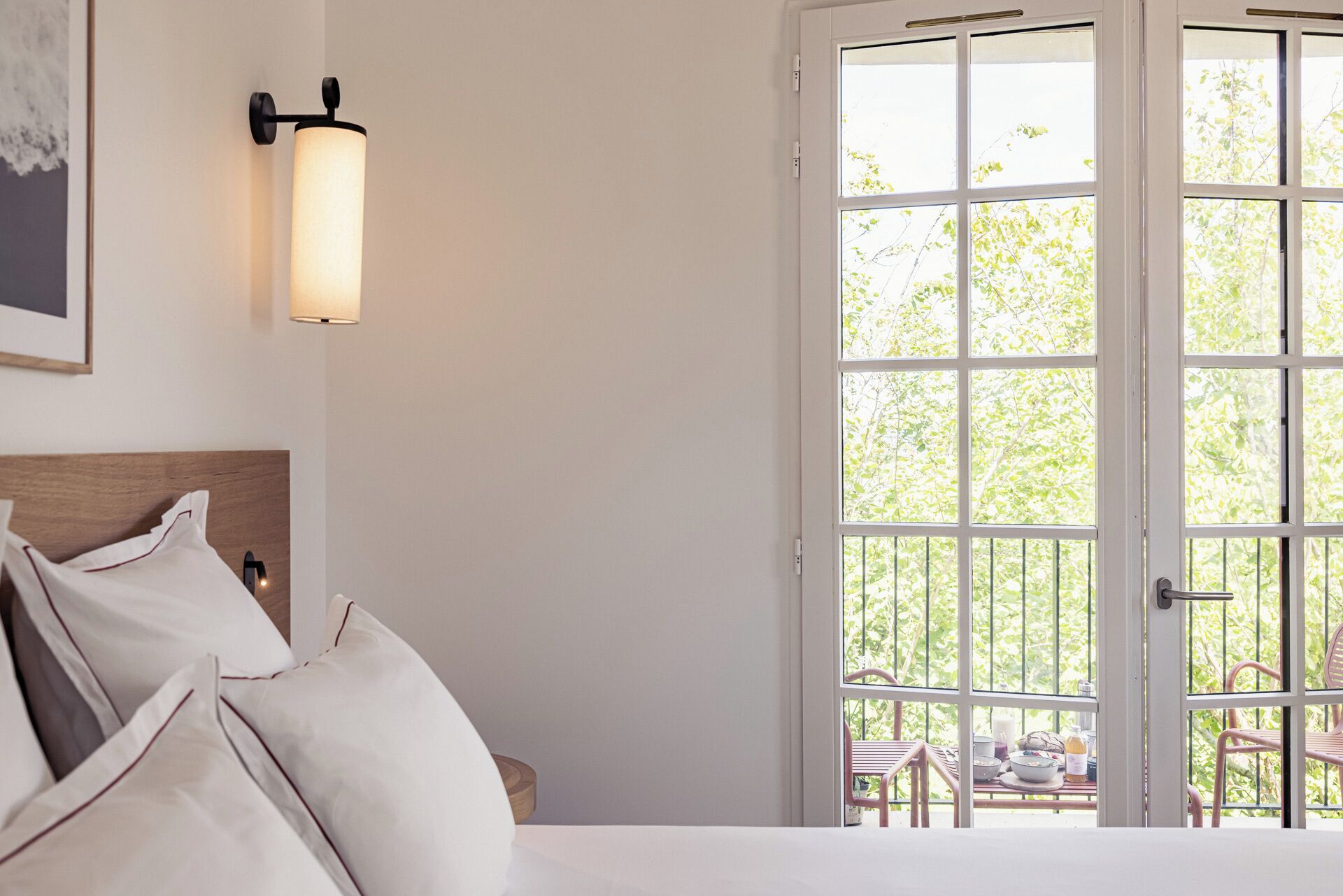A bedroom with a bed, pillows, a bedside table, a wall-mounted light, and a large window with a view of greenery outside.