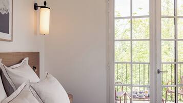A bedroom with a bed, pillows, a bedside table, a wall-mounted light, and a large window with a view of greenery outside.